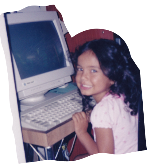 Old photo of a young girl with dark curly hair, smiling over her shoulder at the camera while seated at a beige old computer and keyboard on a wooden desk.